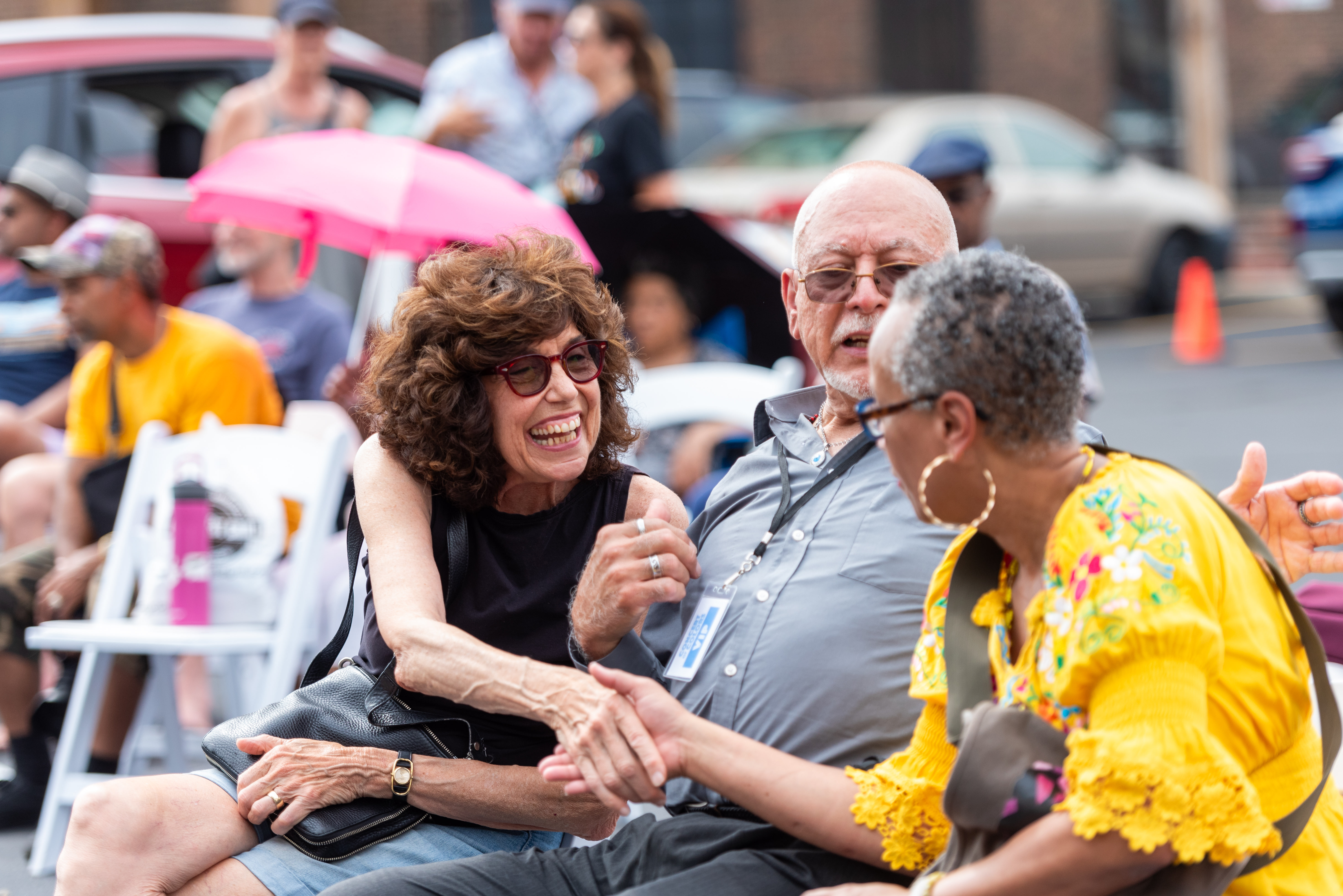 Three people sitting outdoors, smiling and talking. Woman in yellow shakes hands, while others watch with amusement.