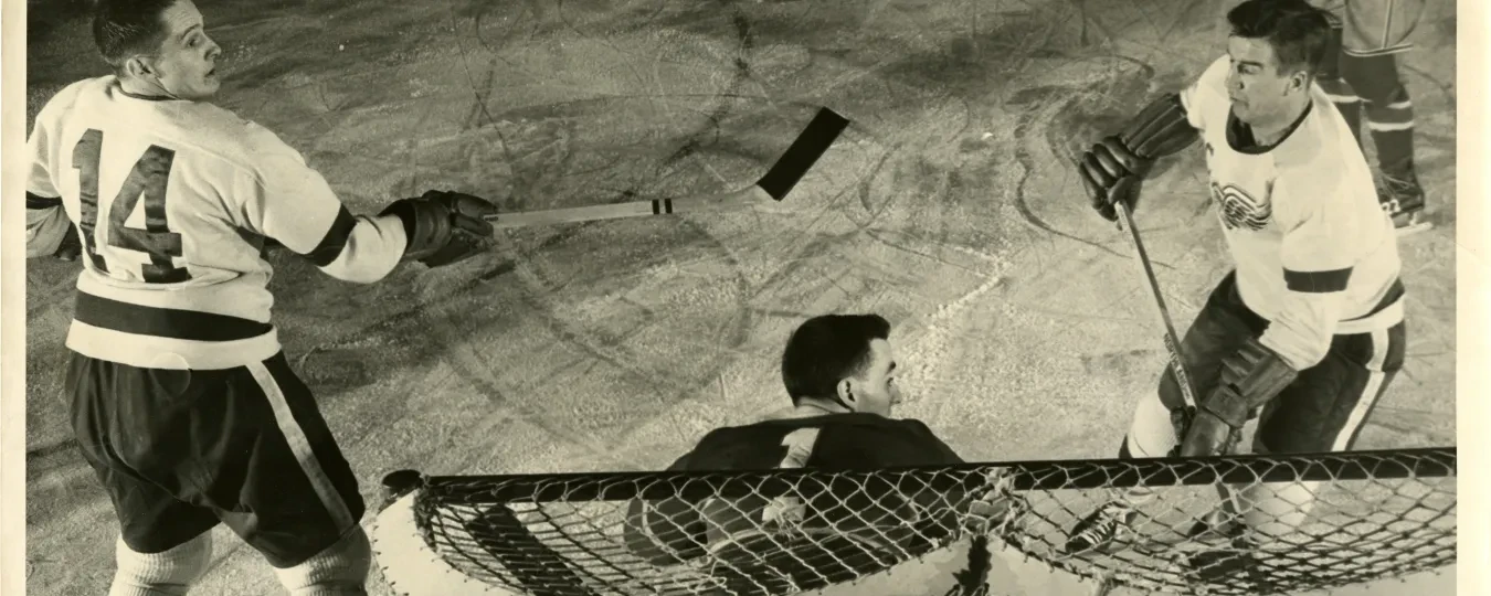 Black and white photo of a hockey game, with two players near the goal and a goalie crouched in position.