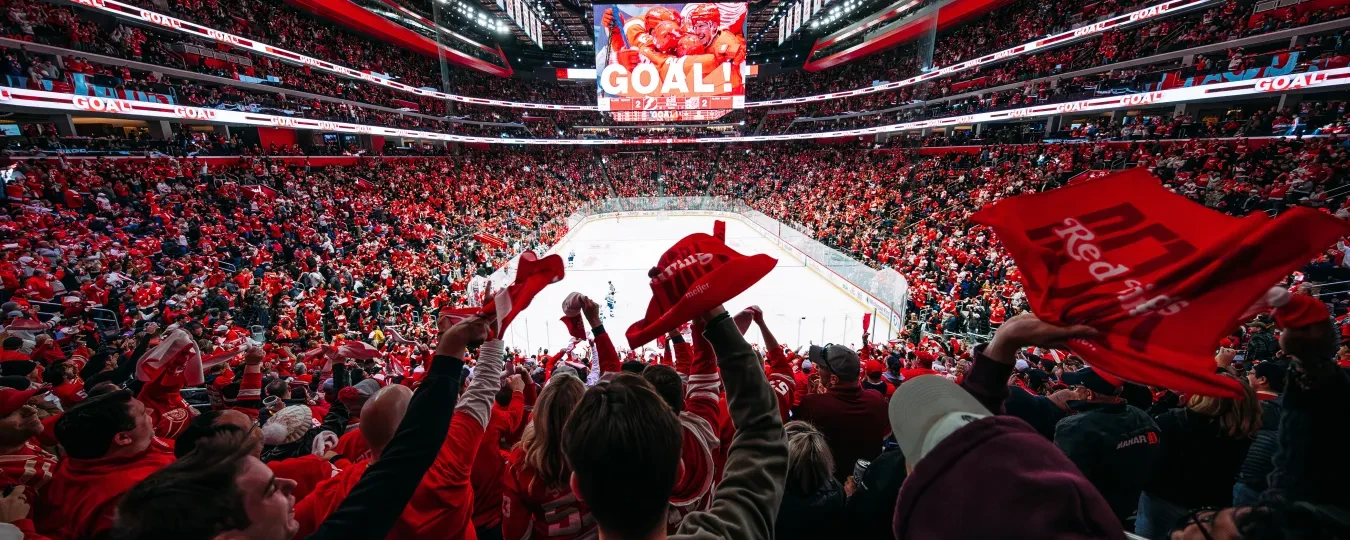 By Allison Farrand. Fans in a stadium cheering a goal, waving red towels, ice rink in the center, and a large screen displaying "Goal!"