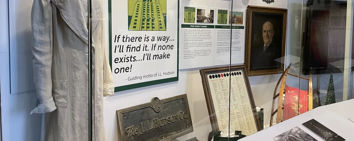 Museum display with historical artifacts, including a dress, framed posters, photographs, and a portrait.