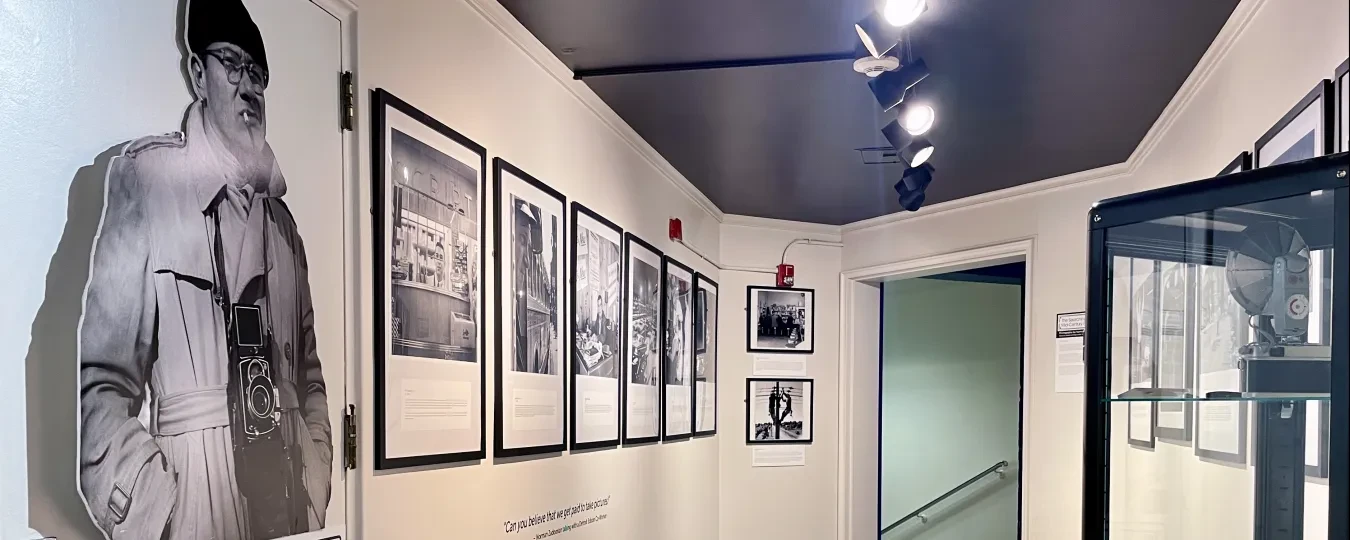 Museum hallway with black-and-white photos on walls, spotlighting a life-sized cutout of a person near glass display case.