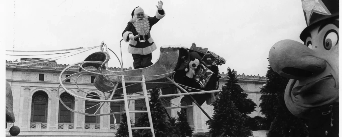 Santa waving from a sleigh float at a parade, with a large toy soldier in the foreground.