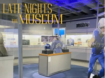 A museum exhibit featuring sports memorabilia from the Detroit Lions, including jerseys, helmets, and a life-size football player cutout in a glass-enclosed space.