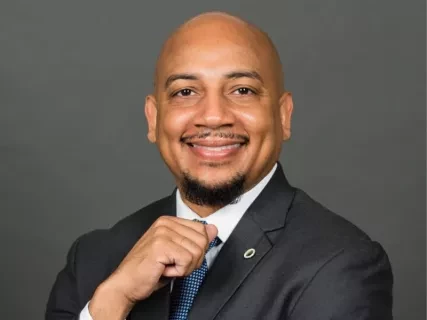 Dr. Ken L. Harris, a smiling Black man in a suit and tie posing with his hand on his chin against a gray background.