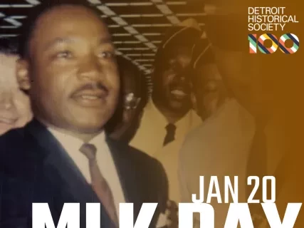 Sepia toned photo of Dr. Martin Luther King Jr wearing a white shirt, dark tie, and dark suit standing in a group of men. January 20 MLK Day Celebration is written in white text over the image.