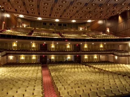Spacious theater interior with rows of empty seats, ornate ceiling, and a red carpeted aisle.