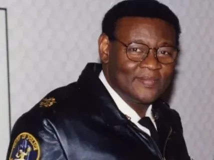 Man in black leather jacket with a badge, wearing glasses and smiling.