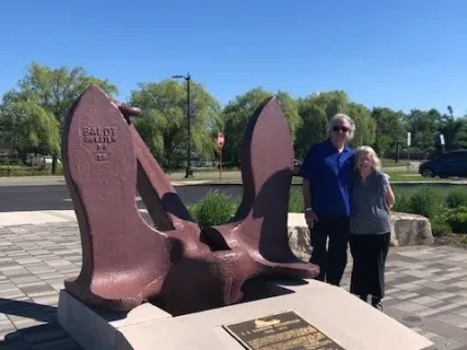 Two people stand beside a large anchor sculpture on a sunny day, with trees and a blue sky in the background.