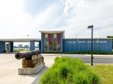Cannon displayed outside a modern museum with a blue facade and large windows, surrounded by greenery.