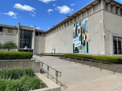 Museum exterior with modern architecture, a large colorful banner, and clear blue sky.