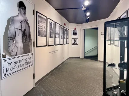 Museum hallway with black-and-white photos on walls, spotlighting a life-sized cutout of a person near glass display case.