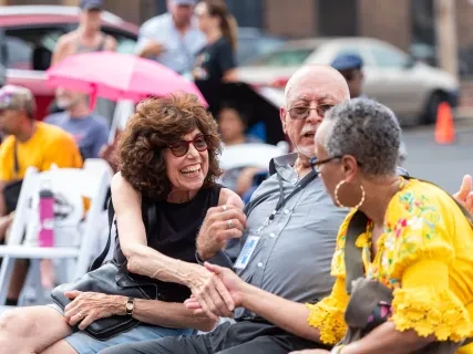 Three people sitting outdoors, smiling and talking. Woman in yellow shakes hands, while others watch with amusement.