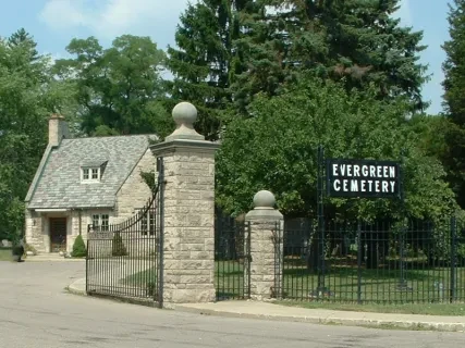 Evergreen Cemetery entrance with stone gates and a small stone building, surrounded by trees.