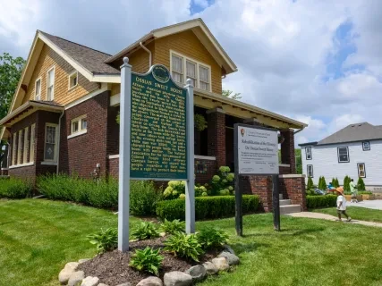 Historic brown house with yellow trim, green sign in front, lush garden, and a partly cloudy sky.