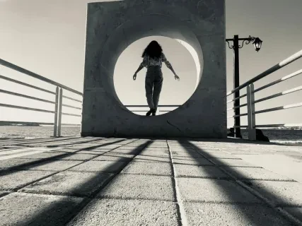 Person standing inside a circular sculpture on a pier, with shadows cast by the railings. Black and white photo.