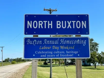 Roadside sign for North Buxton, announces the annual homecoming during Labour Day weekend. Bright blue sky background.