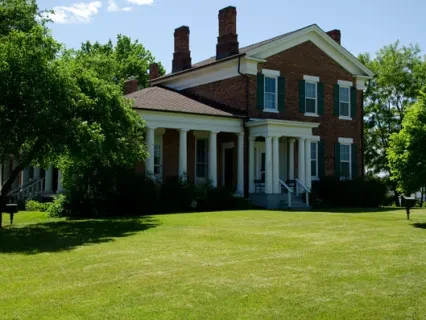 Historic brick house with green shutters, large porch, and lush trees on a sunny day.