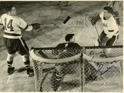 Black and white photo of a hockey goalie guarding the goal as two opposing players approach.