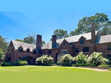 Large brick house with steep roofs, multiple chimneys, and surrounded by lush greenery against a clear blue sky.