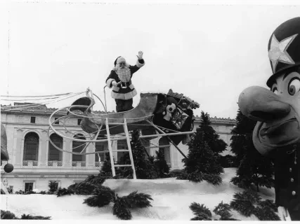 Santa waving from a sleigh float at a parade, with a large toy soldier in the foreground.