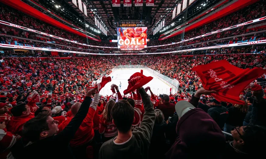 By Allison Farrand. Fans in a stadium cheering a goal, waving red towels, ice rink in the center, and a large screen displaying "Goal!"