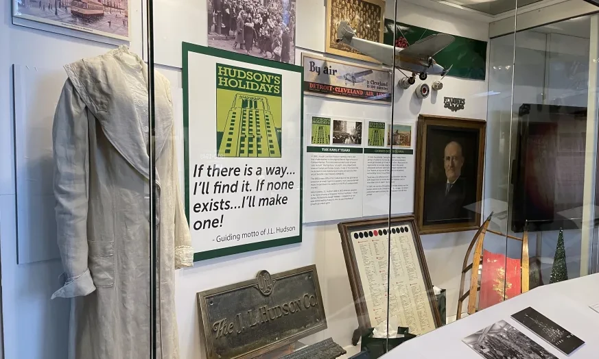 Museum display with historical artifacts, including a dress, framed posters, photographs, and a portrait.