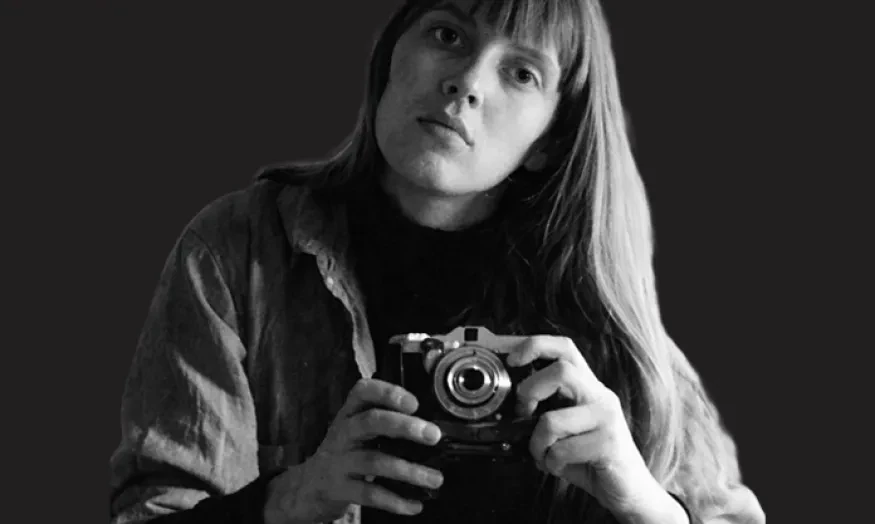 Black and white photo of a person with long hair holding a camera against a dark background.