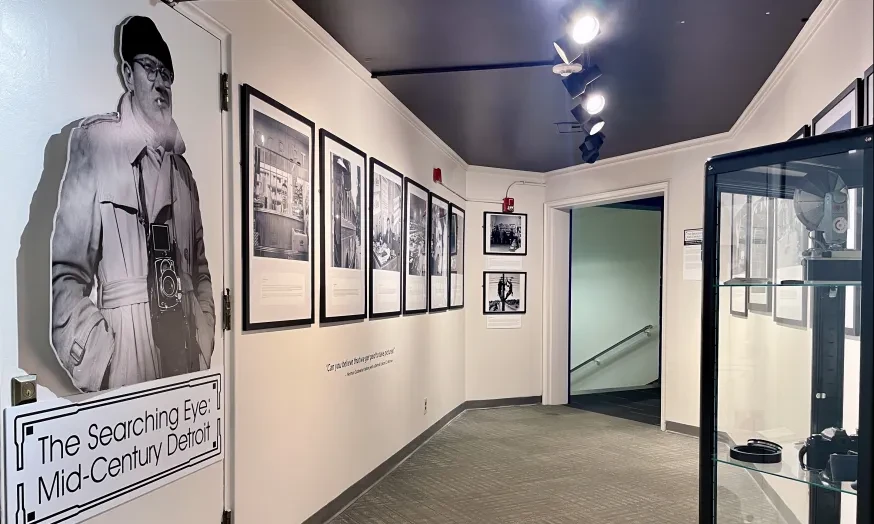 Museum hallway with black-and-white photos on walls, spotlighting a life-sized cutout of a person near glass display case.