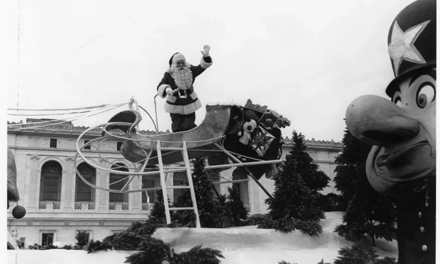 Santa waving from a sleigh float at a parade, with a large toy soldier in the foreground.