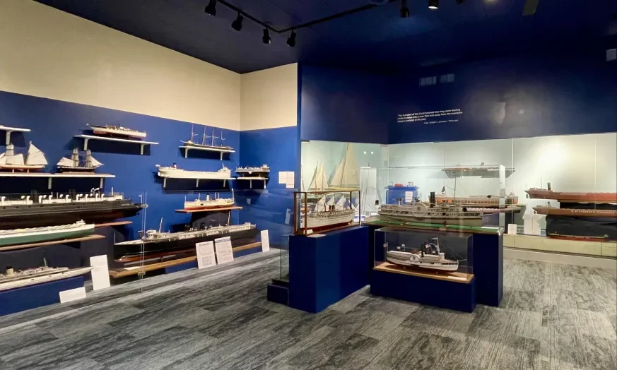 Museum exhibit room with model ships displayed on walls and in glass cases, under dim lighting.