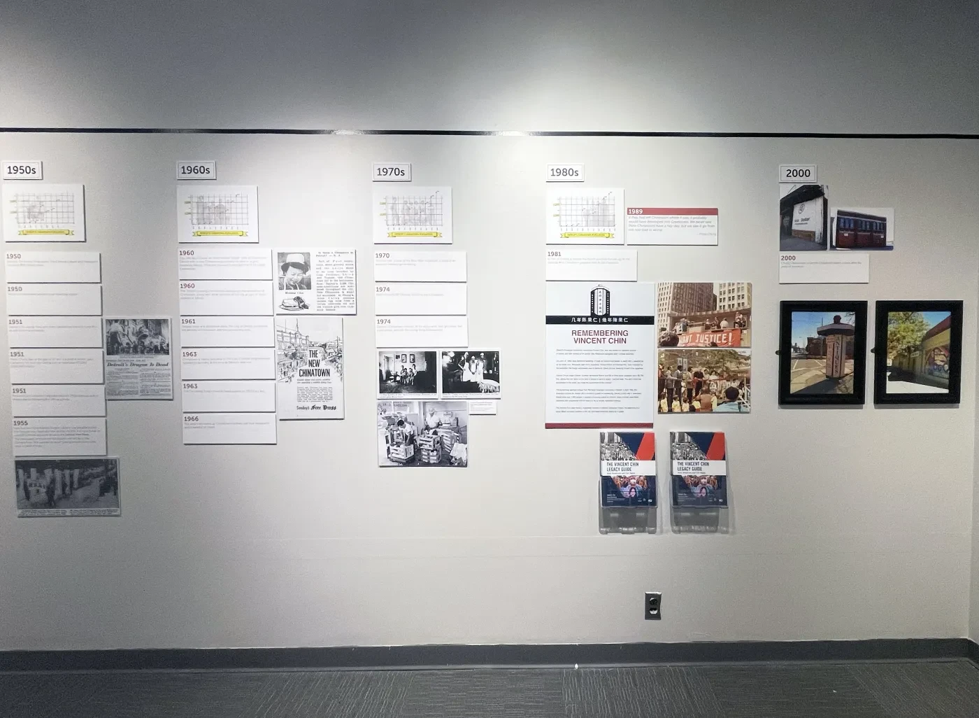 Exhibit wall with documents, black-and-white photos, and framed pictures arranged in rows on a light-colored background.