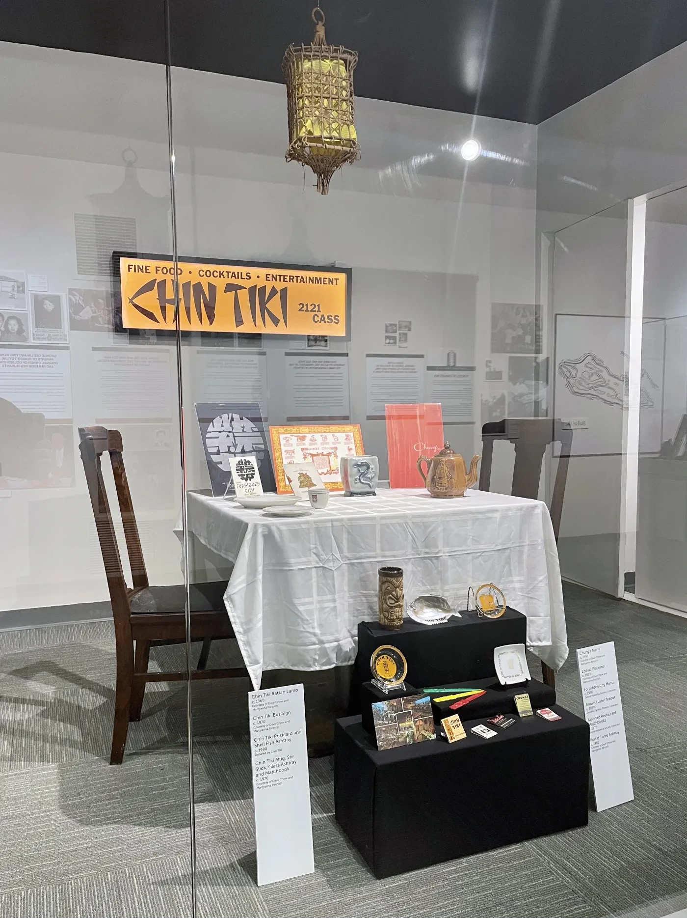 Exhibit with dining table, chairs, lantern, and vintage items on display, featuring a sign for "Chan Tiki" in a glass case.