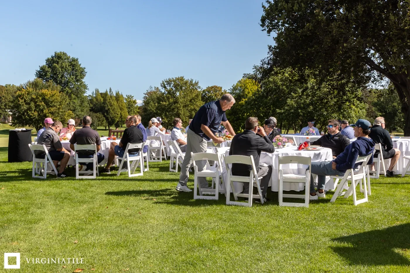 People seated at round tables on a lawn, under clear blue skies, surrounded by trees, during a sunny outdoor gathering.