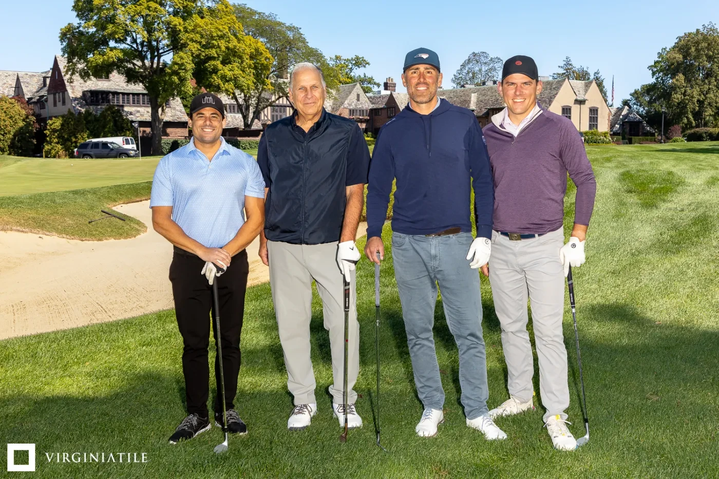Four men smiling on a golf course, each holding a club, with trees and a clubhouse in the background.