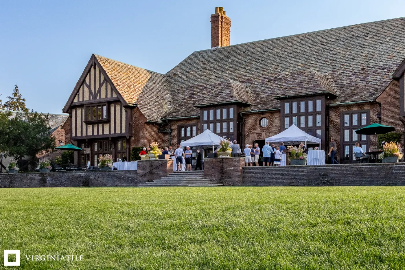 A Tudor-style building with a sloped roof, people gathered outside, green lawn in foreground, and blue sky above.