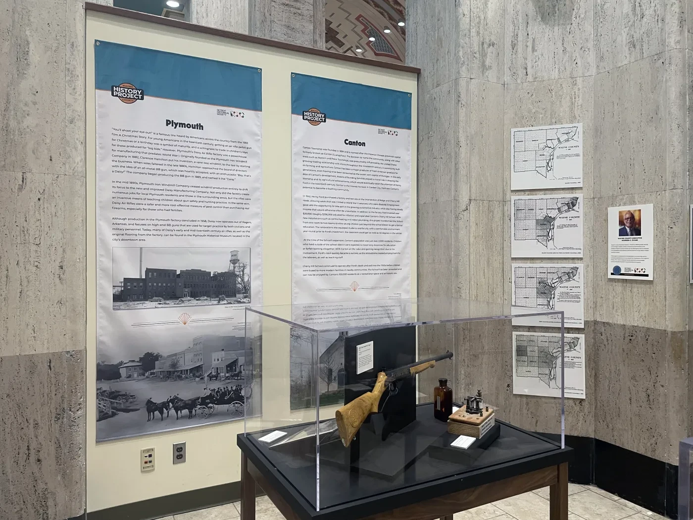Museum exhibit with historical photos, documents, and artifacts enclosed in a glass display case against a marble wall.