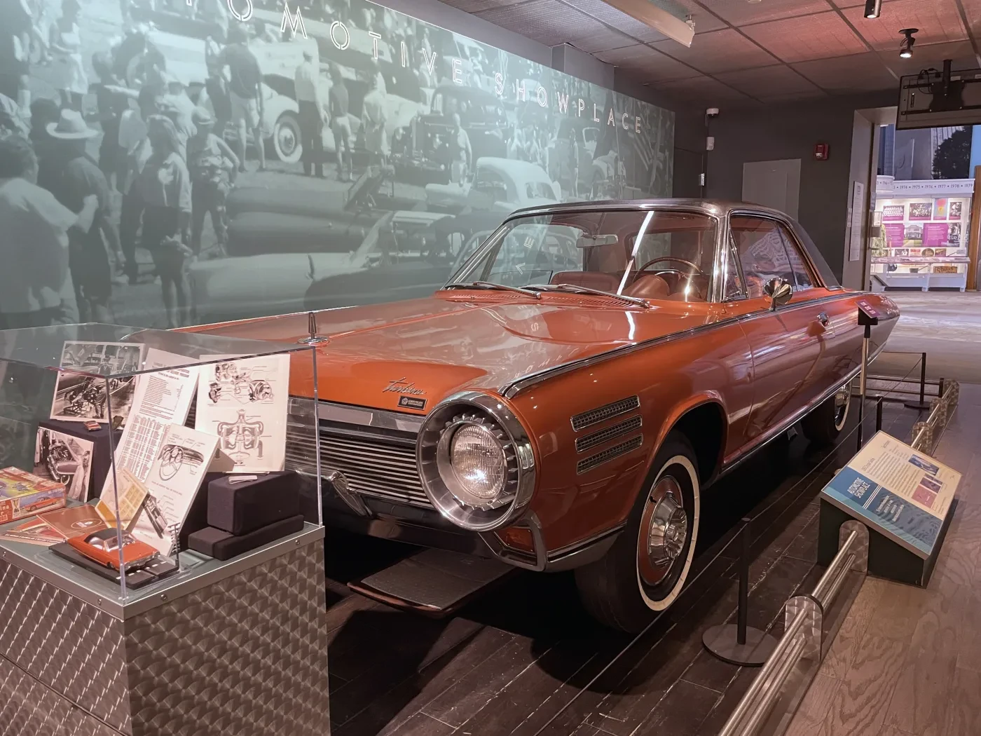 Red vintage car displayed in a museum, surrounded by informational panels and a black-and-white wall mural.