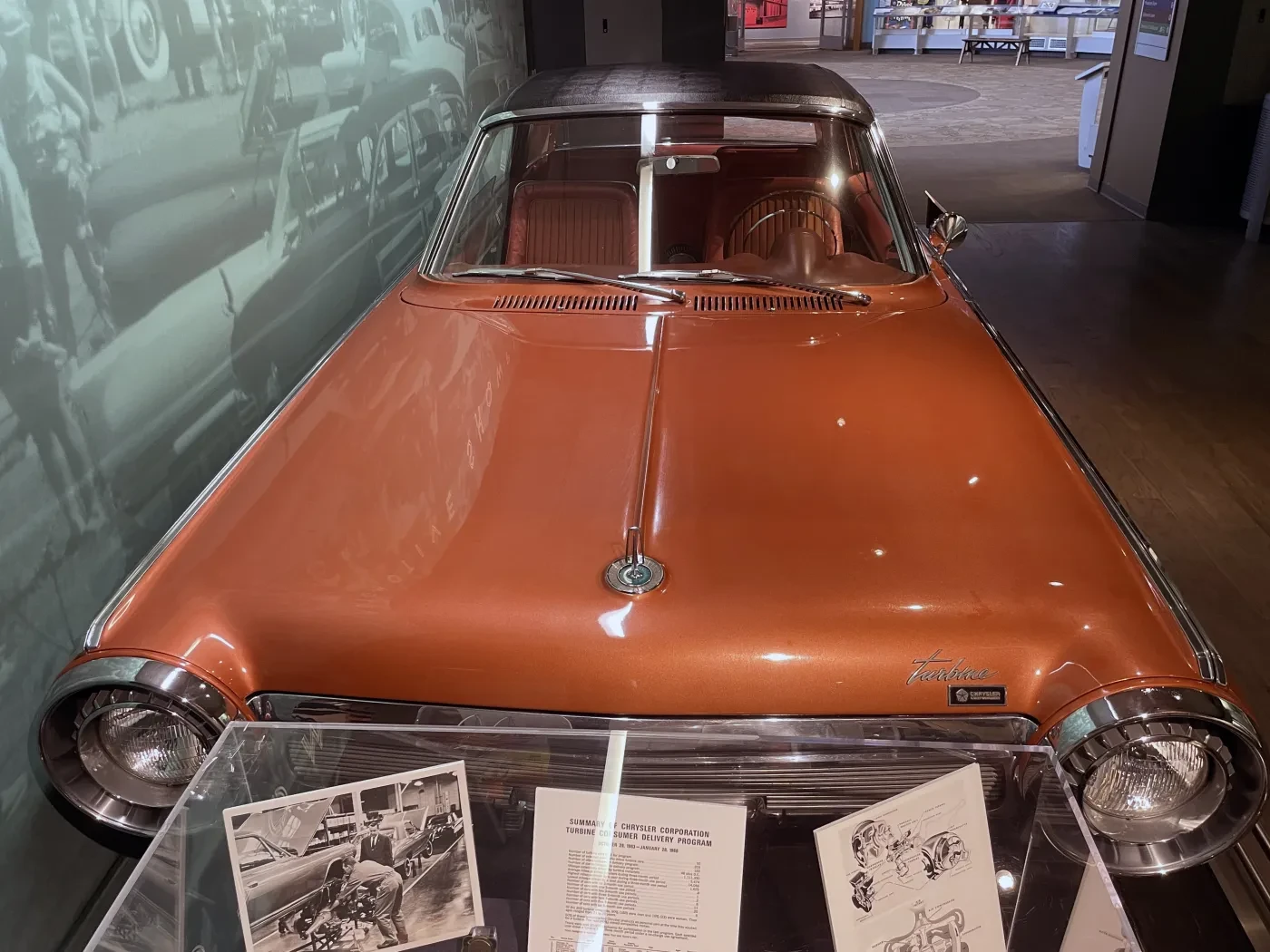 Vintage orange car displayed in a museum, with historical photos and sketches on a nearby desk.