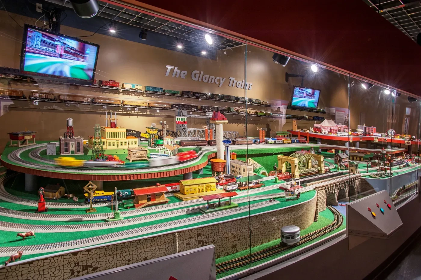 Model train exhibit with intricate tracks, colorful trains, and miniature buildings, under soft lighting in a display case.