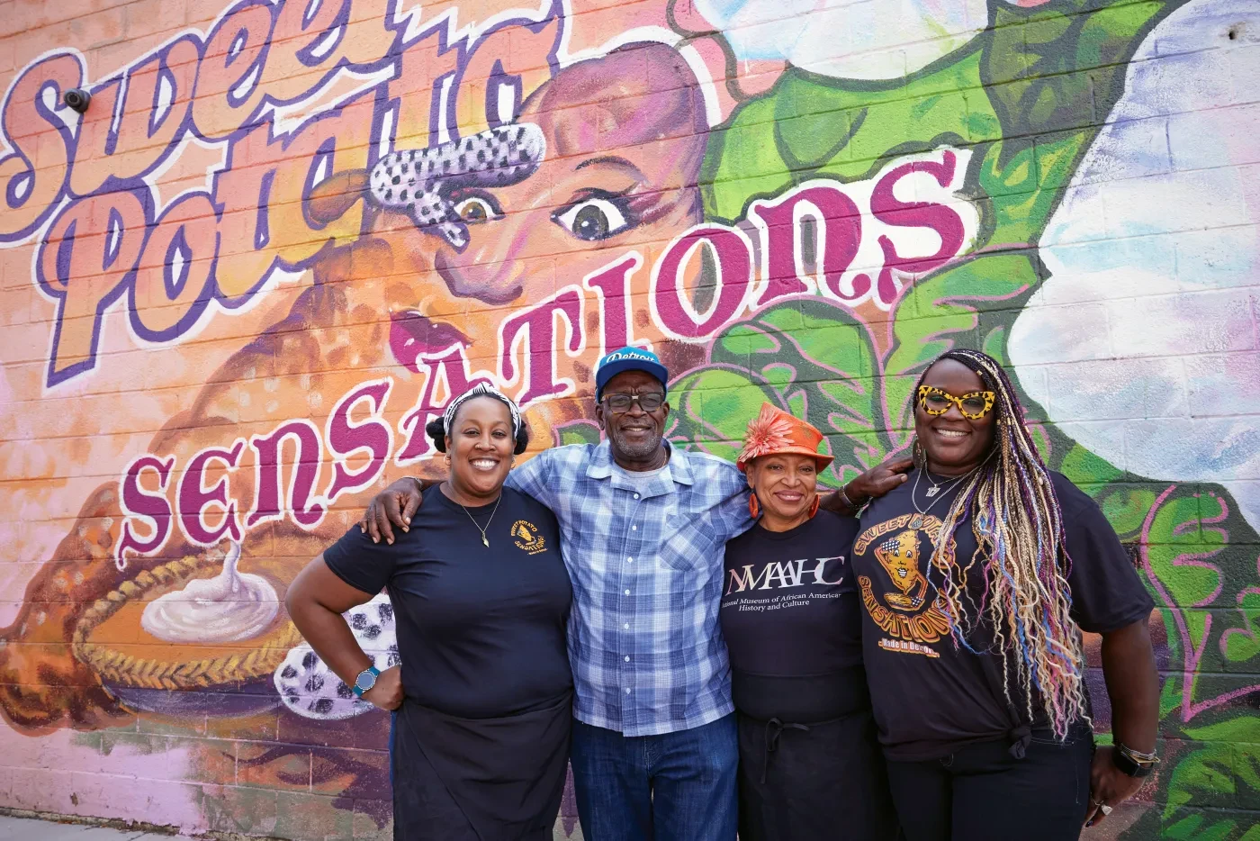 Four people smiling in front of a colorful mural reading "Sweet Potato Sensations."