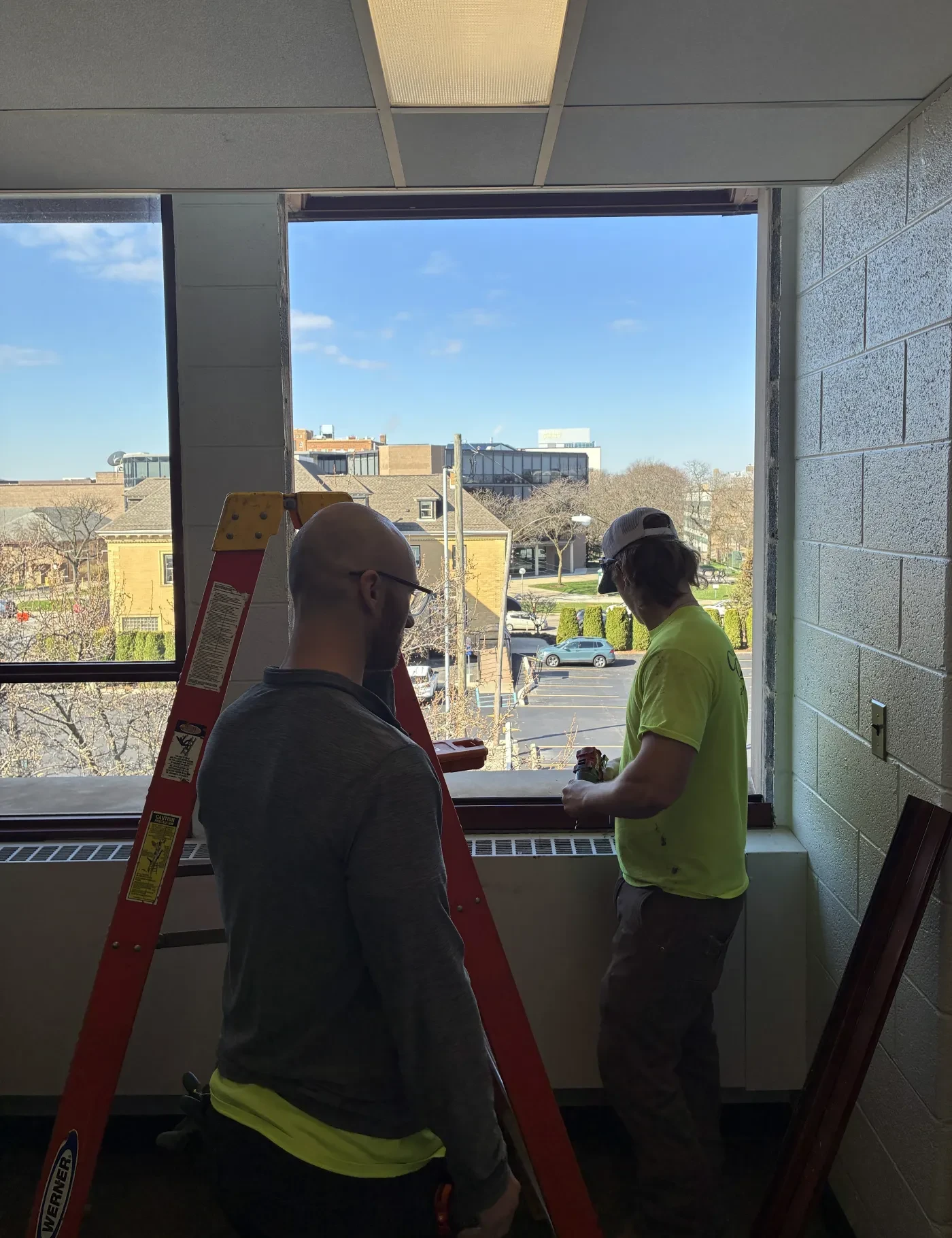 Two men working by an open window, one standing on a ladder. Bright day outside with buildings in view.