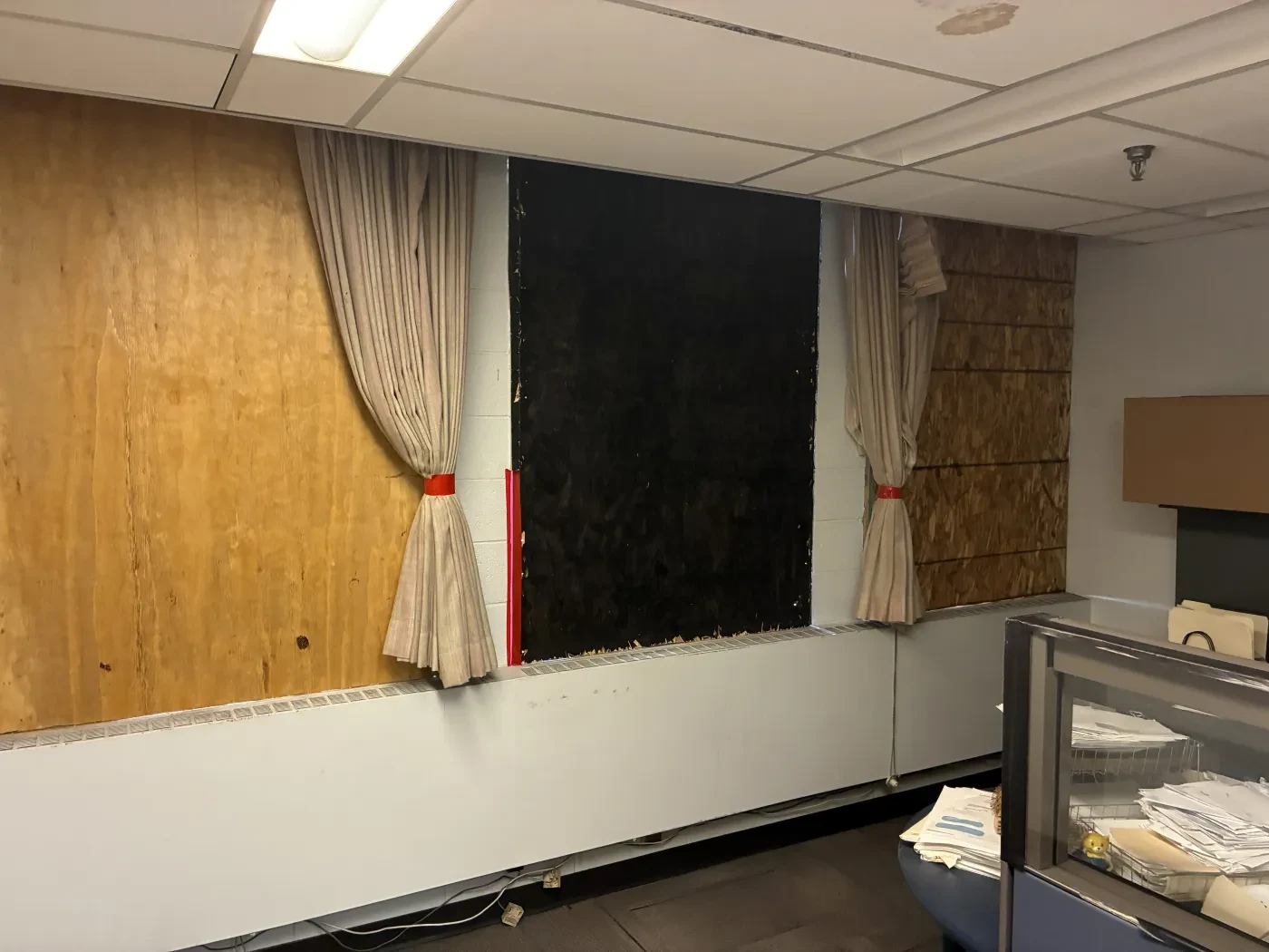 Office windows covered with plywood, curtains tied back. Ceiling lights and office furniture visible.