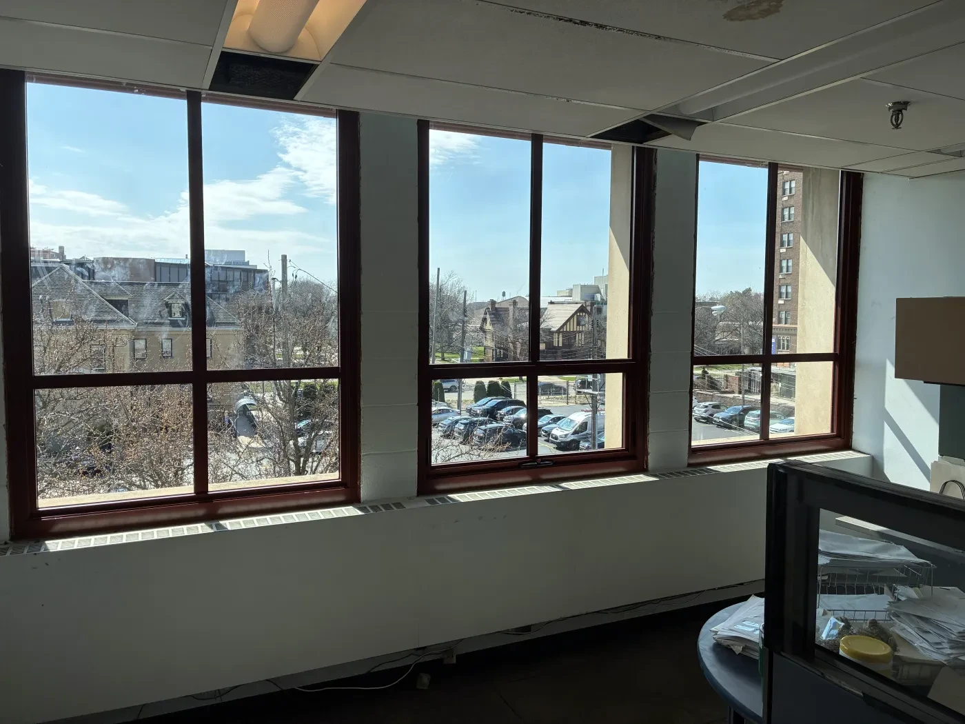Sunlit room with large windows, overlooking trees and a parking lot.