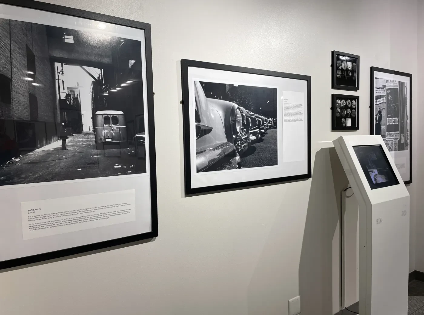 Black and white photographs displayed in a gallery, with a digital kiosk nearby.
