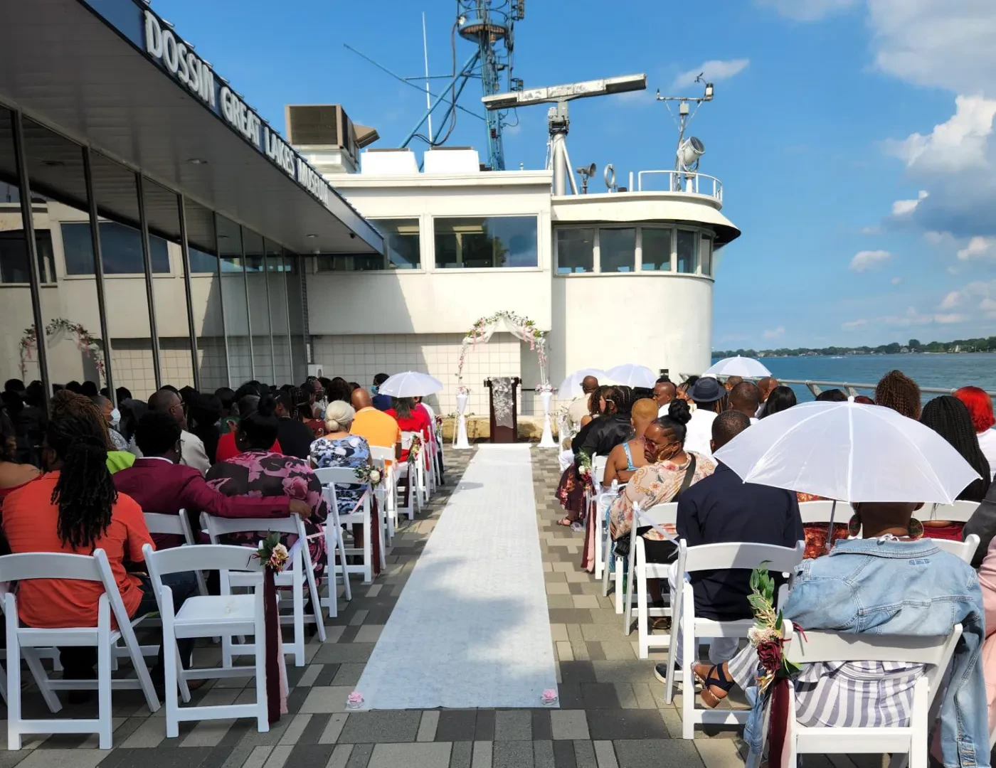 Outdoor wedding ceremony on a boat deck with guests seated under umbrellas on a sunny day.