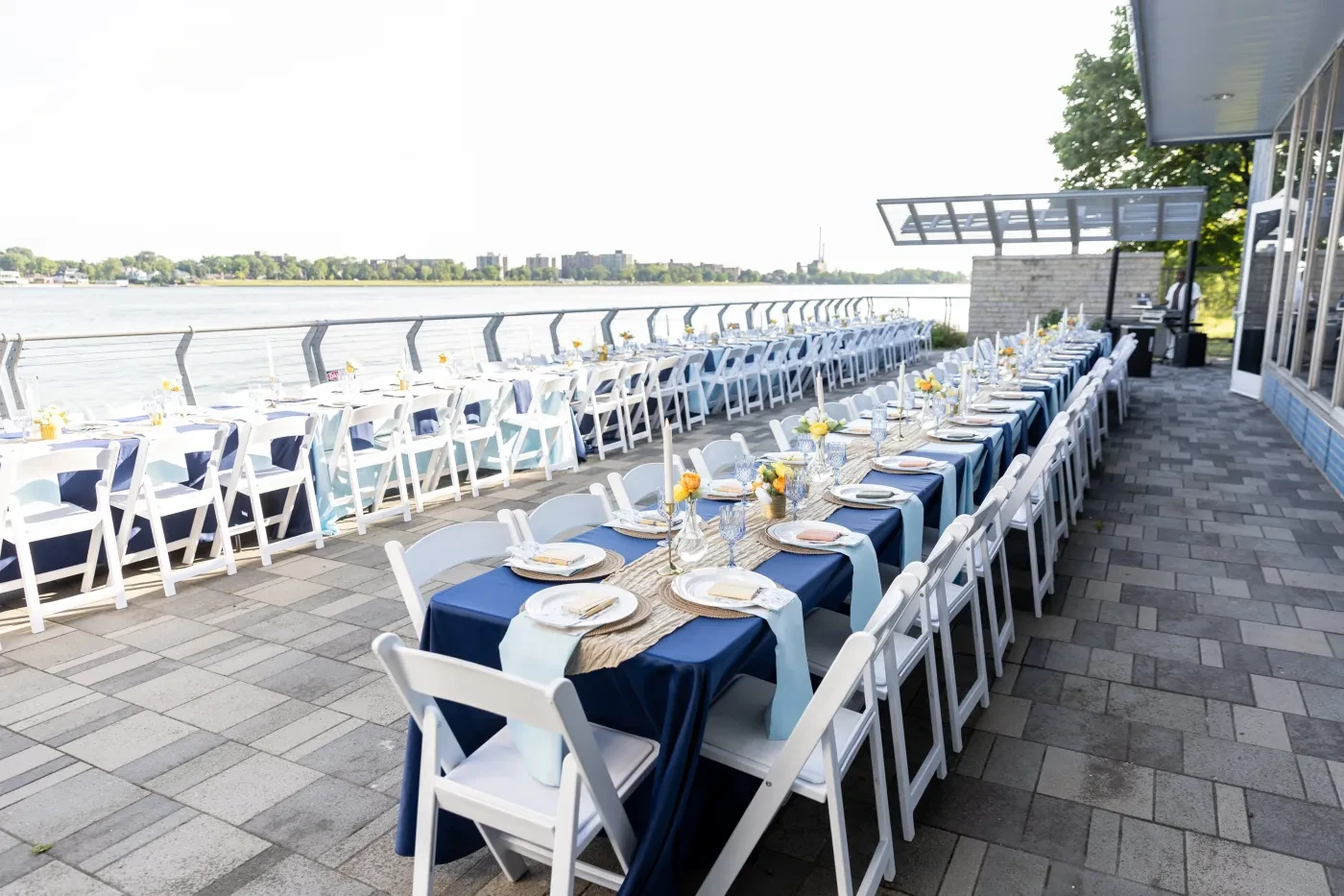 Outdoor dining setting with long tables, blue tablecloths, white chairs, overlooking a body of water.
