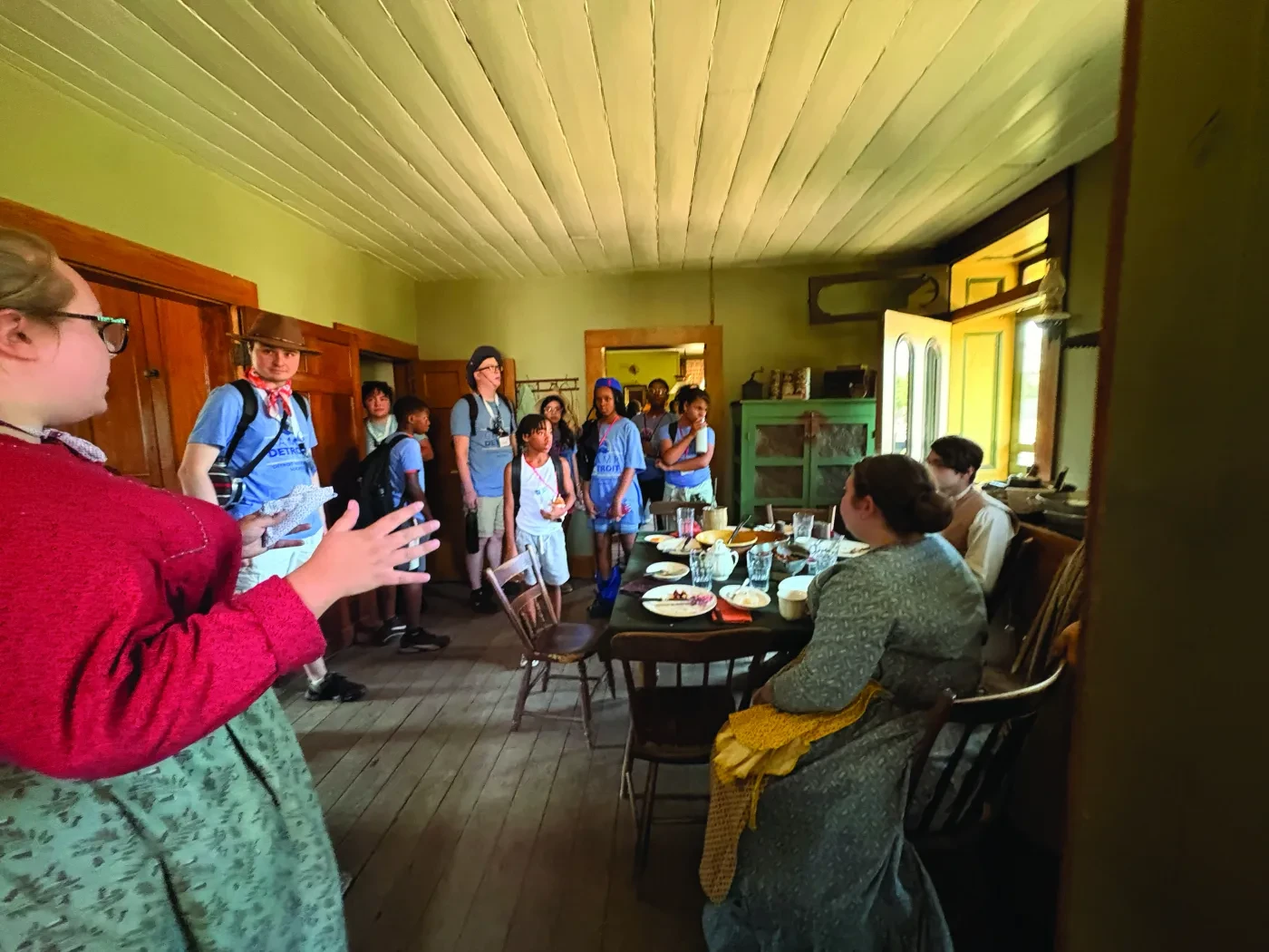 Group in historical attire gathered in a rustic dining room with a wooden table set for a meal.