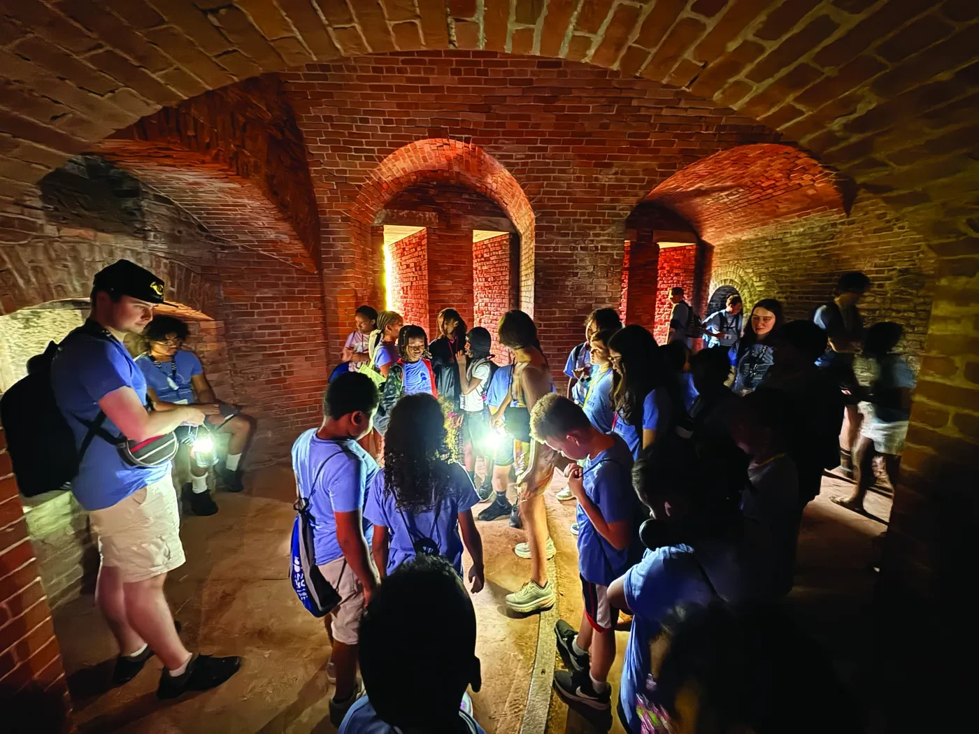 Group of children with flashlights exploring a dimly lit brick-arched tunnel.