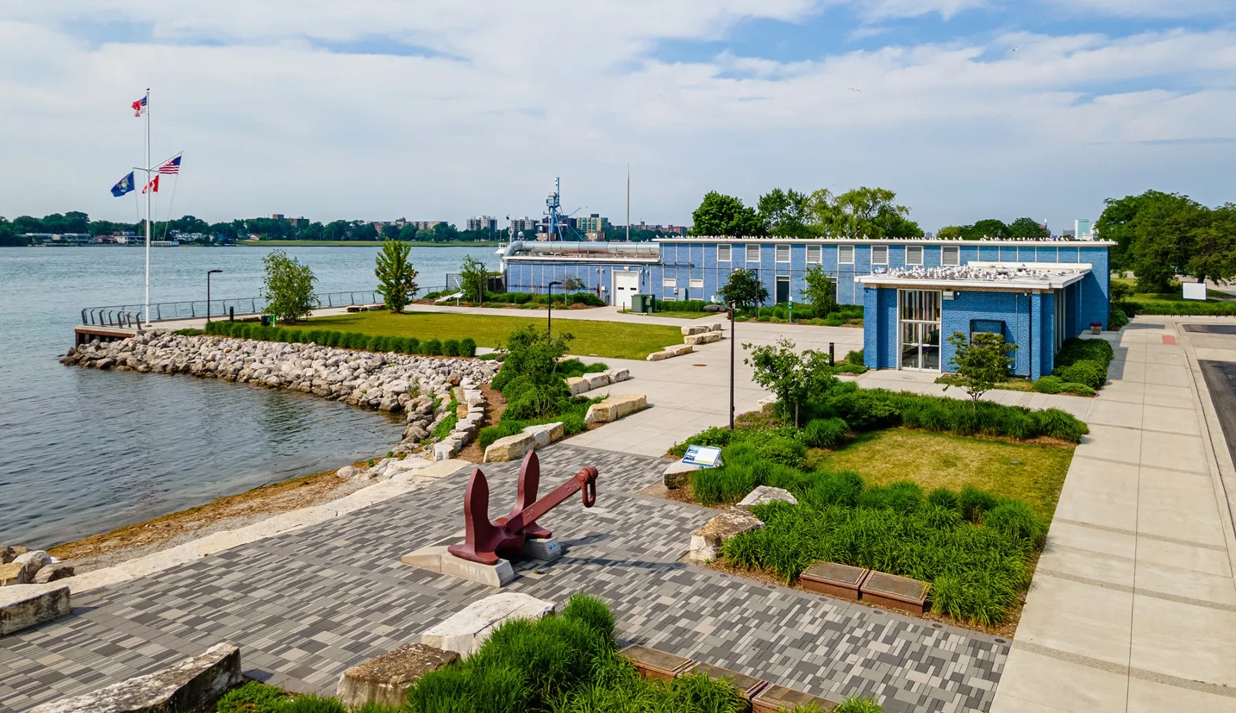 Waterfront park with stone path, flags, a large anchor sculpture, and a modern building. Trees and water in the background.