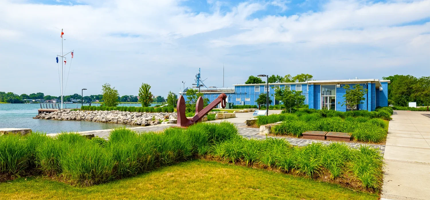 Lakeside park with green lawns, a flagpole, an anchor sculpture, and a blue building under a partly cloudy sky.
